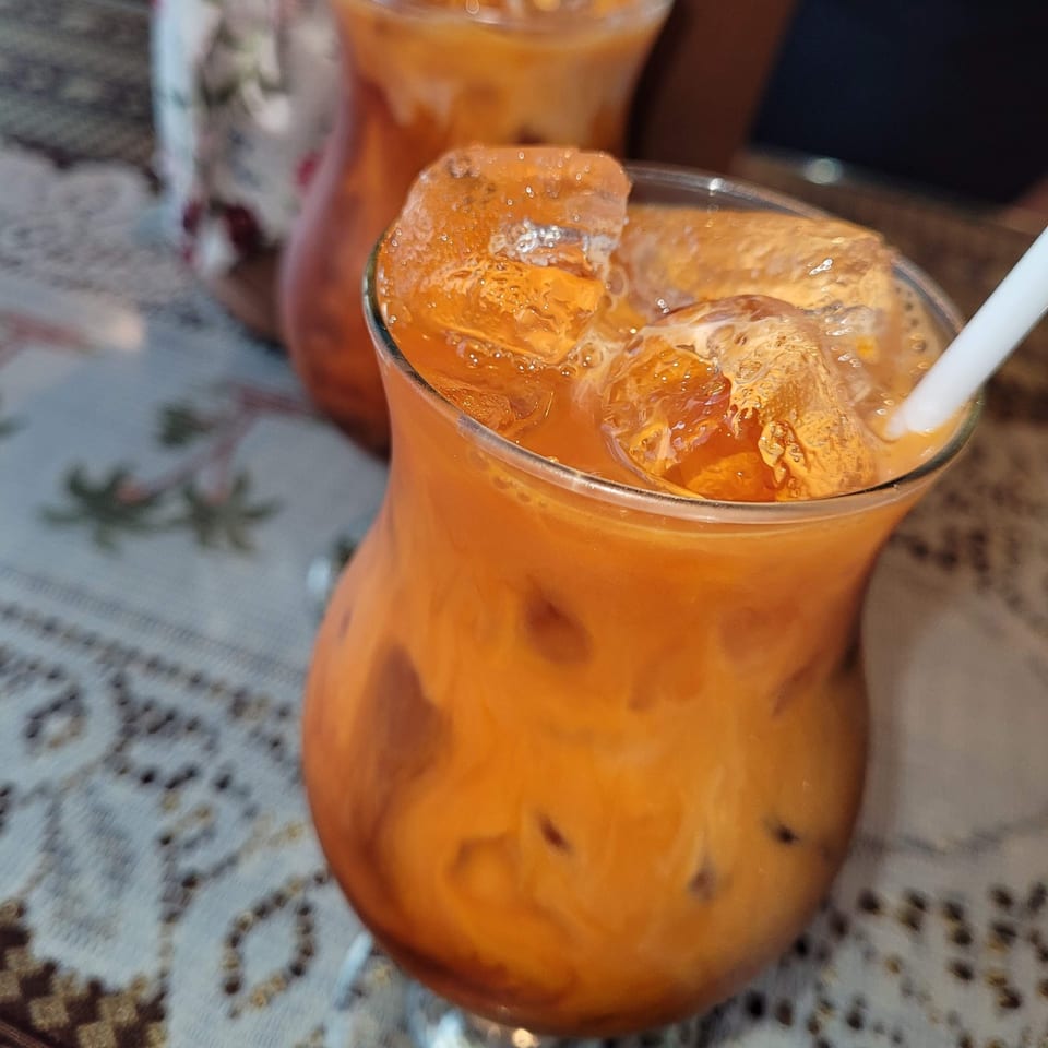 Two glasses of iced Thai tea with a white straw on a patterned tablecloth.