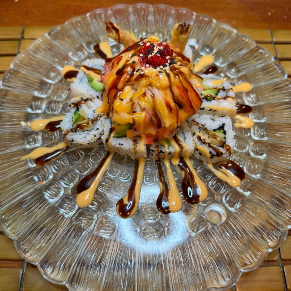 Sushi rolls topped with shrimp, salmon, avocado, sesame seeds, and colorful sauces on a decorative glass plate.